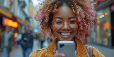 Smiling woman with curly pink hair and orange glasses, wearing a yellow jacket, looking at her smartphone while walking down a busy street.