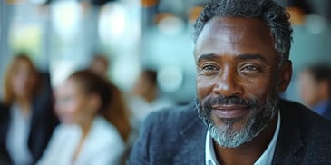 Smiling man with a salt-and-pepper beard and hair in a dark blazer, sitting in a professional setting with blurred people in the background.