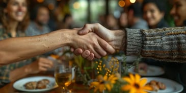 Two people shaking hands over a dining table with yellow flowers, surrounded by smiling people enjoying a meal in a warmly lit setting.