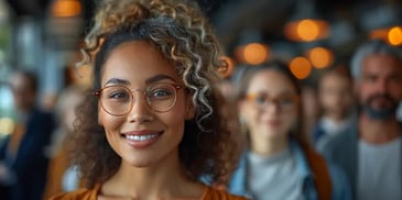 Smiling woman with curly hair and glasses, wearing an orange top, standing in a crowd with warm lighting in the background.