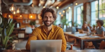 Smiling man with wavy hair and a yellow shirt, sitting at a laptop.