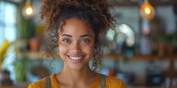 Smiling woman with curly hair wearing a mustard yellow top and overalls, standing in a warmly lit, cozy indoor setting.