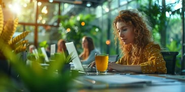 Focused woman with curly hair working on a laptop in a green, sunlit office with a glass of juice nearby.