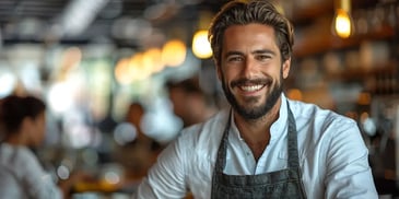 Smiling man with a beard, wearing an apron and white shirt, standing in a restaurant setting.