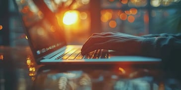 Hands typing on a laptop keyboard with a sunset and bokeh lights in the background.