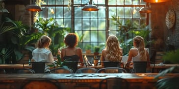 Four women with laptops work together at a large wooden table in a sunlit, plant-filled room with large windows.