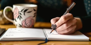 Woman writing in notebook with floral mug.