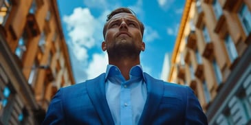 Confident man in a blue suit standing between tall buildings, looking upward at the blue sky.