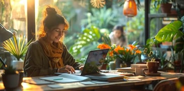 Smiling woman with a bun and mustard scarf working on a laptop at a wooden desk.